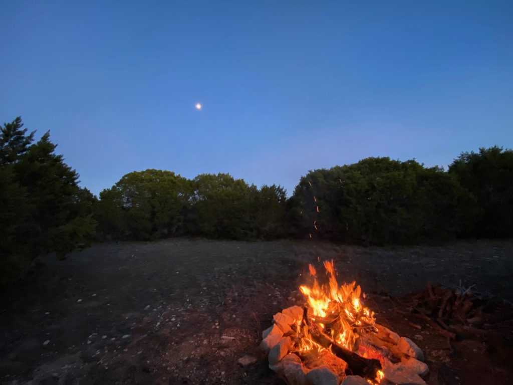 Campfire burning in a stone fire ring at dusk, with trees behind.